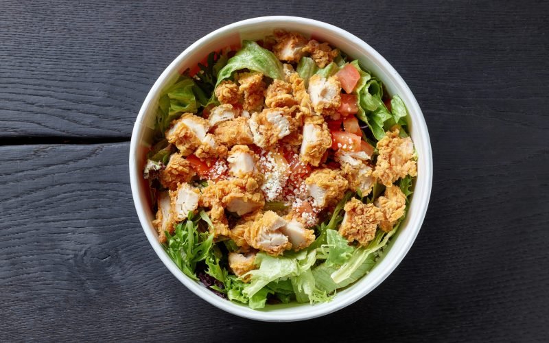 fresh caesar salad with fried chicken meat in paper take away container on black wooden table, top view