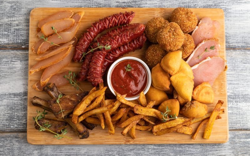 set of snacks: sausages, fried potatoes, meat balls, dumplings, basturma on a cutting board on a gray wooden background. Top view, flat lay, close up.